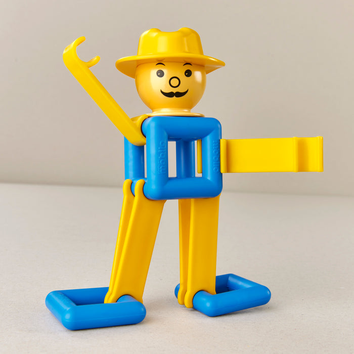 A small plastic toy figure in bright yellow and blue, posed with one arm raised and a rectangular piece extending from the side, on a plain light background.