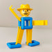 A small plastic toy figure in bright yellow and blue, posed with one arm raised and a rectangular piece extending from the side, on a plain light background.