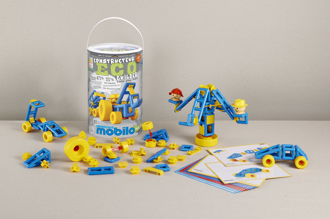 A toy construction set displayed on a tabletop with its cylindrical packaging bucket. The set includes blue and yellow plastic building blocks, small vehicles, instruction cards, and a crane model built from the pieces.