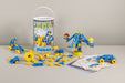 A toy construction set displayed on a tabletop with its cylindrical packaging bucket. The set includes blue and yellow plastic building blocks, small vehicles, instruction cards, and a crane model built from the pieces.