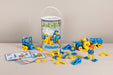 A Mobilo Construction Builder toy set displayed with its clear storage bucket, scattered blue and yellow building pieces, and instruction sheets on a light beige surface against a neutral beige background.