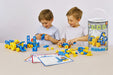 Two children playing with blue and yellow mobile construction blocks on a light beige table, with a tall branded Mobilo storage bucket and instruction sheets nearby against a plain light gray studio background.