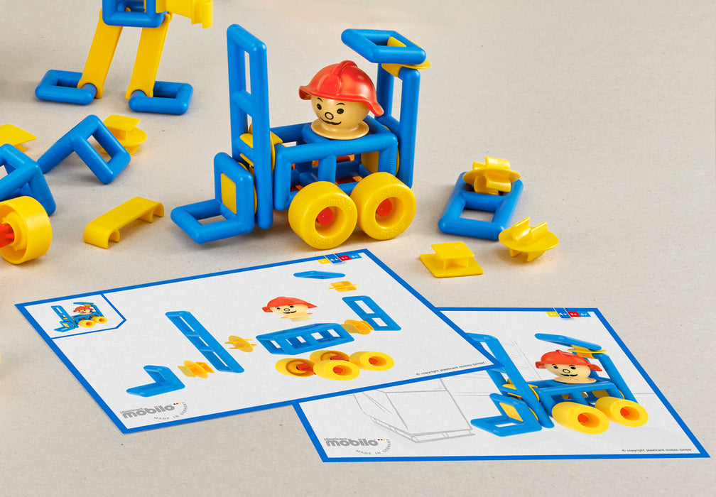 A set of blue and yellow modular building blocks with a small minifigure, arranged on a light beige surface alongside two printed instruction sheets. The instruction sheets show step-by-step assembly diagrams with the same colorful pieces and a minifigure wearing a red hard hat.